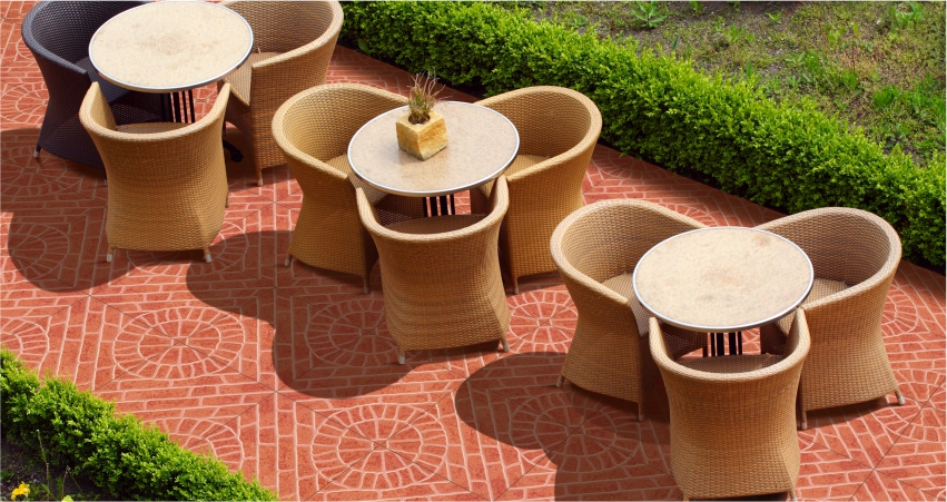 An outdoor patio with red patterned tiles, wicker chairs, round tables, and green hedges